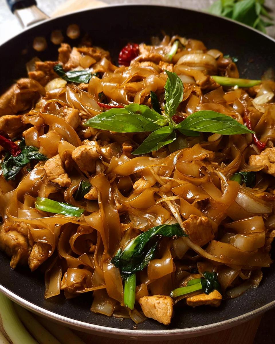 Close-up of Street Style Thai Drunken Noodles with chicken, wide rice noodles, and basil in a black pan.