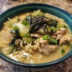 A close-up of a bowl of Creamy Garlic Chicken Ramen, featuring noodles, chicken pieces, bok choy, and nori.