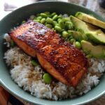 A close-up of a Crispy Salmon and Rice Bowl featuring a perfectly cooked salmon fillet, fluffy white rice, edamame, and sliced avocado.