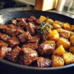 Close-up of juicy garlic steak bites and golden roasted potatoes in a skillet, garnished with rosemary.