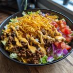 A delicious High-Protein Cheeseburger Bowl with seasoned ground beef, shredded cheese, lettuce, tomatoes, and red onion, drizzled with sauce.