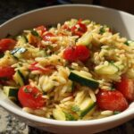 A bowl of lemony orzo salad with fresh veggies, featuring cherry tomatoes, cucumber, and herbs.