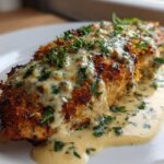 A close-up of a golden-brown Parmesan Crusted Chicken breast generously topped with creamy garlic sauce and fresh parsley.