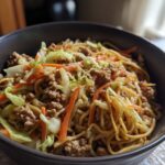 A close-up of a dark bowl filled with a Potsticker Noodle Bowl featuring pork, noodles, shredded cabbage, and carrots.