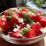 Close-up of a clear bowl filled with cubed watermelon, crumbled feta cheese, and fresh mint leaves, perfect for watermelon recipes.